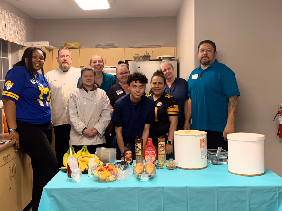 A group of nine people standing behind a table covered with a blue tablecloth in a kitchen setting. The table has two large ice cream containers, various toppings, bananas, and bowls of fruit. The people are smiling and appear to be staff or participants in an event.