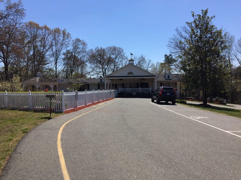 Front entrance of a single-story senior living building with a driveway, white picket fence, gazebo, and a parked car.