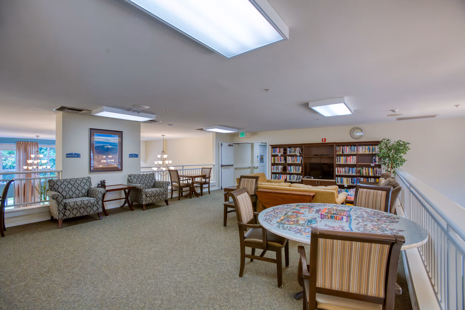 A spacious, well-lit common area in an assisted living facility featuring a round table with chairs, a seating area with armchairs and a couch, bookshelves filled with books, and a large TV. The room has carpeted floors, overhead fluorescent lighting, and a railing overlooking a lower level with large windows and hanging chandeliers.