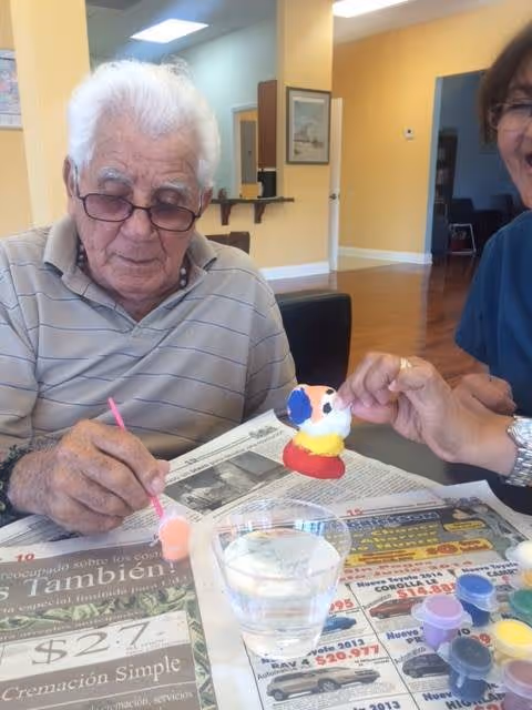 An elderly man wearing glasses and a striped polo shirt is painting a small figurine with a pink paintbrush while sitting at a table covered with newspapers. A woman next to him is holding the figurine steady. There are small paint containers and a glass of water on the table. The background shows a room with yellow walls and wooden flooring.