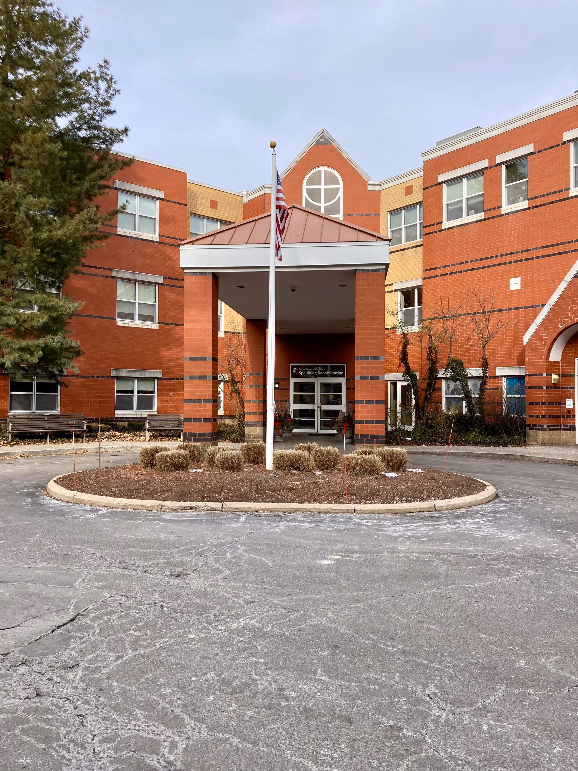Front entrance of Spaulding Nursing & Therapy Center showing a red brick building with a covered driveway, an American flag on a flagpole in the center, and some landscaping with bushes around the driveway circle.
