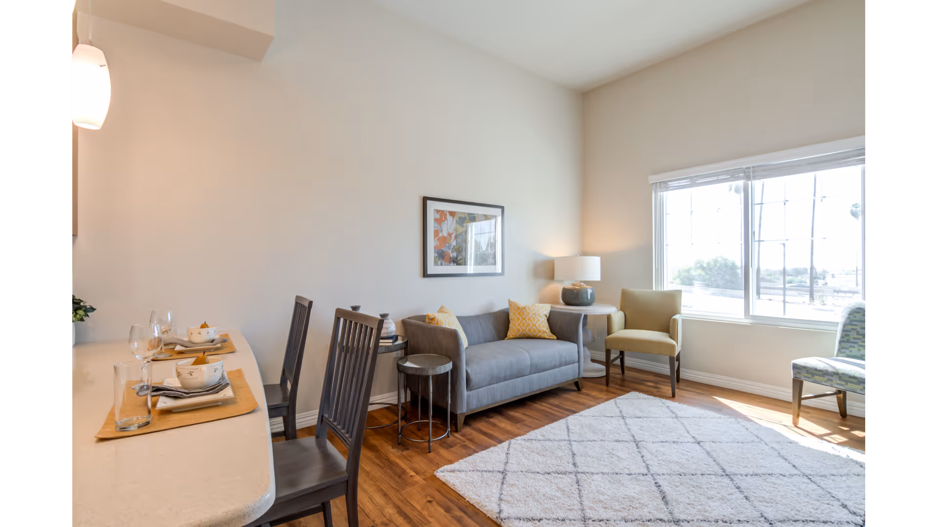 Bright living room with a gray sofa, two chairs, a patterned rug, and a breakfast counter set with place settings beside a large window.