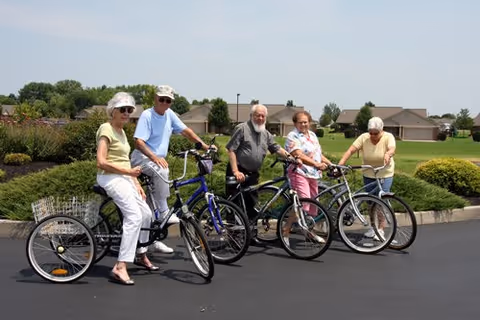 Five elderly individuals standing outdoors on a paved area with their bicycles. They are smiling and appear to be enjoying a sunny day. In the background, there are green bushes, grass, and residential houses under a clear sky.