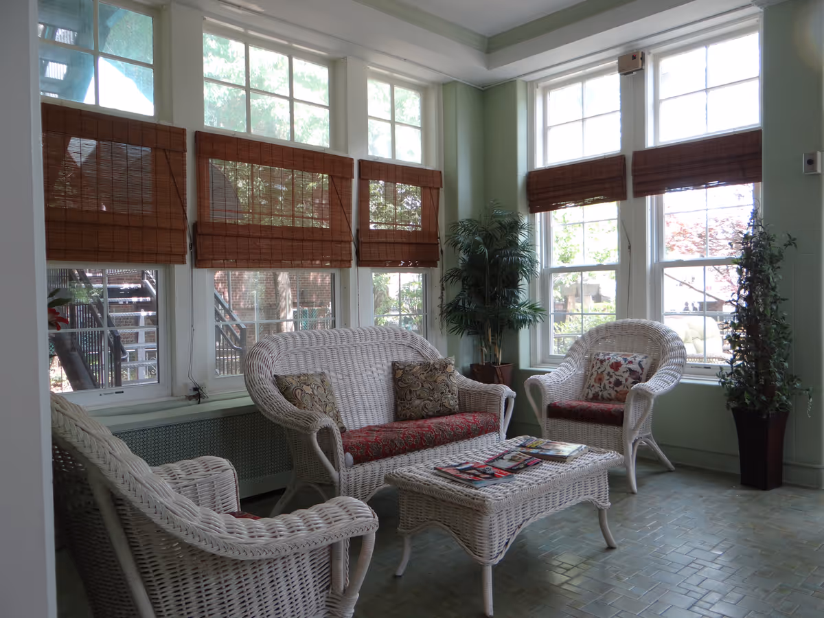A bright sitting area with white wicker furniture including a loveseat, two armchairs, and a coffee table. The furniture has red patterned cushions and decorative pillows. Large windows with brown bamboo blinds allow natural light to fill the room. There are two tall potted plants in the corners, and magazines are placed on the coffee table.