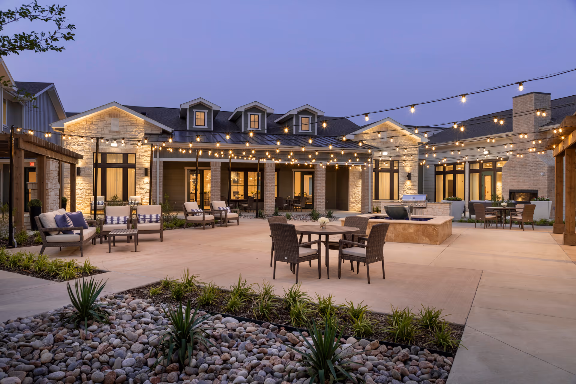 Outdoor patio area at dusk with string lights hanging overhead, multiple seating arrangements including cushioned chairs and tables, a fire pit, and a stone building with large windows and a fireplace in the background.