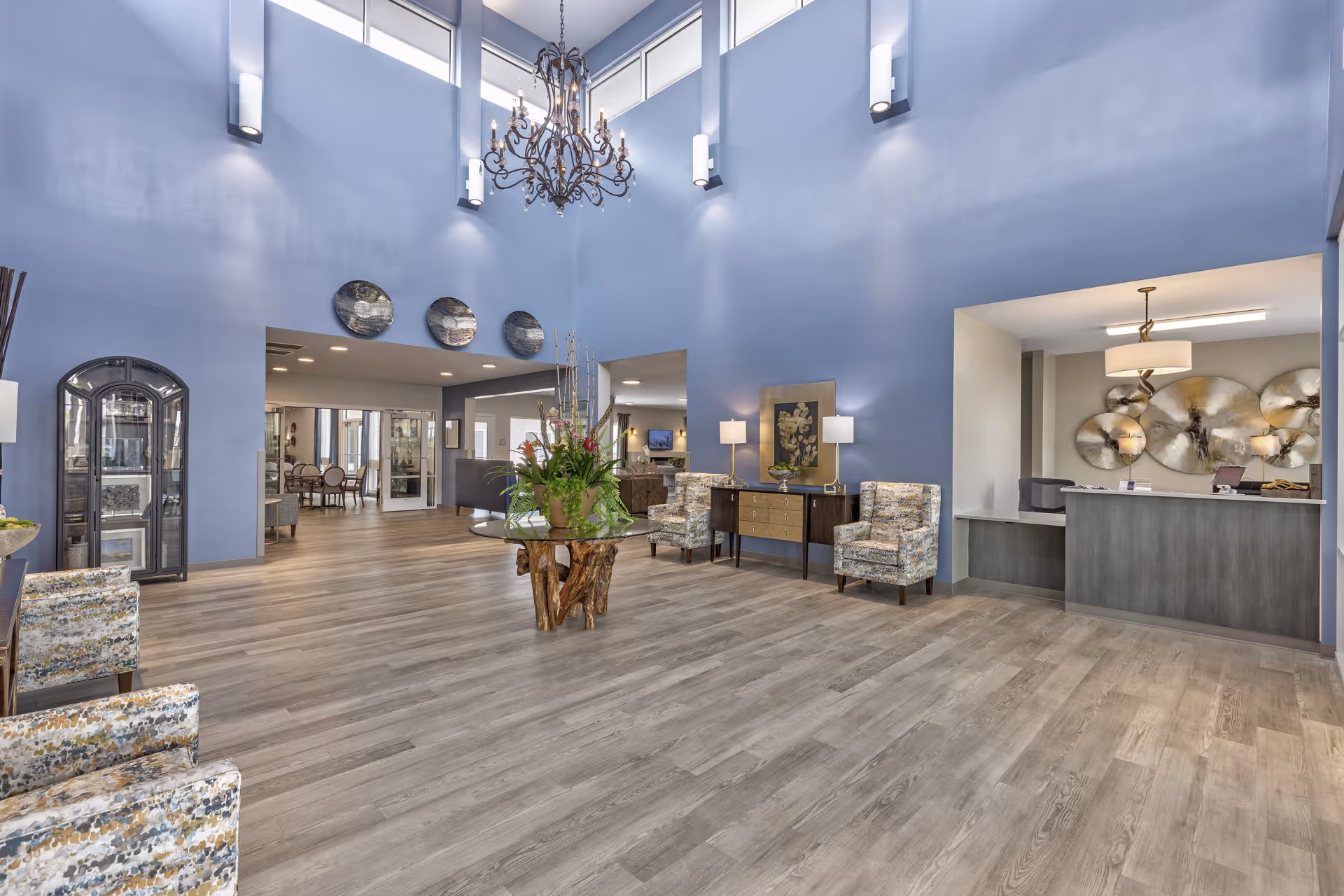 Spacious and well-lit senior living facility lobby with high ceilings painted in light blue, a large chandelier, decorative plates on the wall, a round glass table with a floral arrangement, patterned armchairs, a wooden sideboard with lamps, and a reception desk with modern decor.