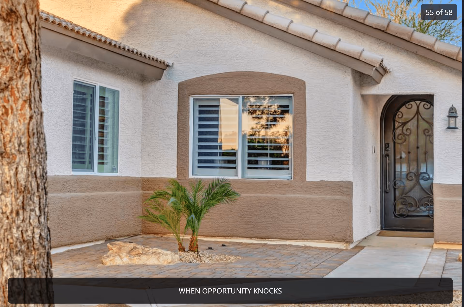 Exterior view of a single-story building with beige and white stucco walls, a black wrought iron front door, two windows with white shutters, a small palm plant, and a paved walkway and patio area.