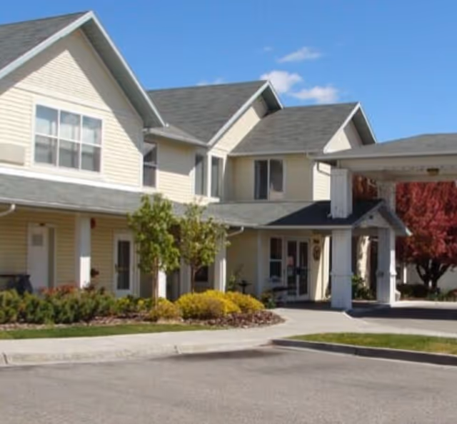 Exterior view of a two-story assisted living facility building with beige siding, multiple windows, a covered entrance, and landscaped greenery including small trees and bushes under a clear blue sky.