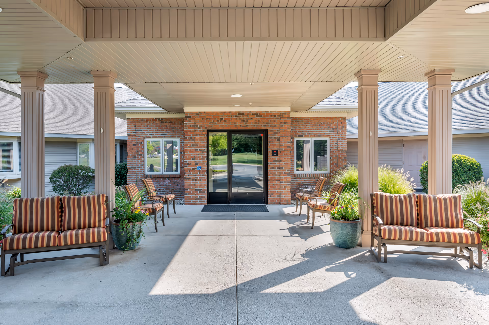 Covered entrance area of a building with a brick facade and glass double doors. There are striped cushioned benches and chairs arranged on both sides, along with large potted plants. The area is shaded by a roof supported by columns, and greenery is visible around the building.
