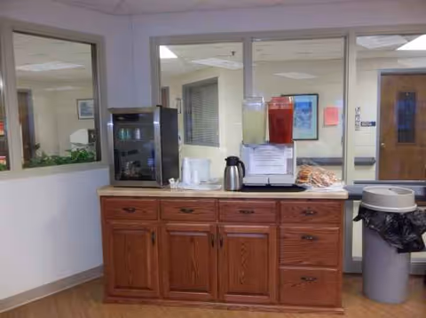 Wooden cabinet counter with coffee machine, beverage dispenser, and pastries at Pine Crest Nursing Home.