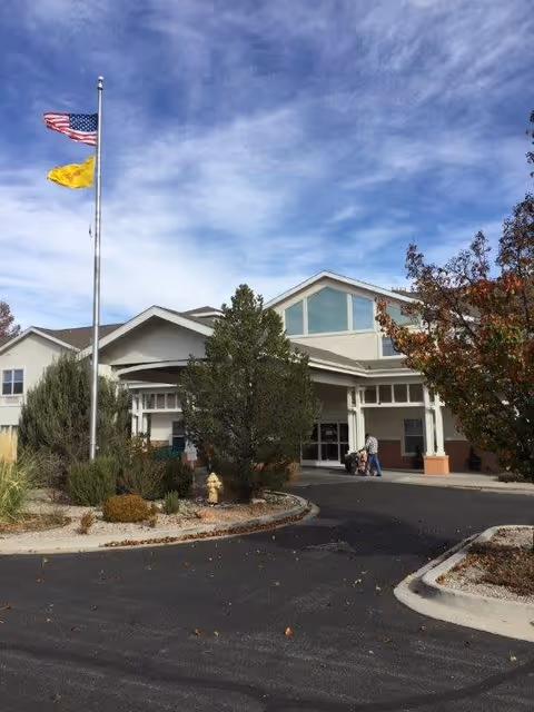Exterior view of Avamere at Rio Rancho facility showing the main entrance with a covered drop-off area, two flagpoles with the American flag and a yellow flag, landscaped bushes and trees, and a person walking near the entrance.