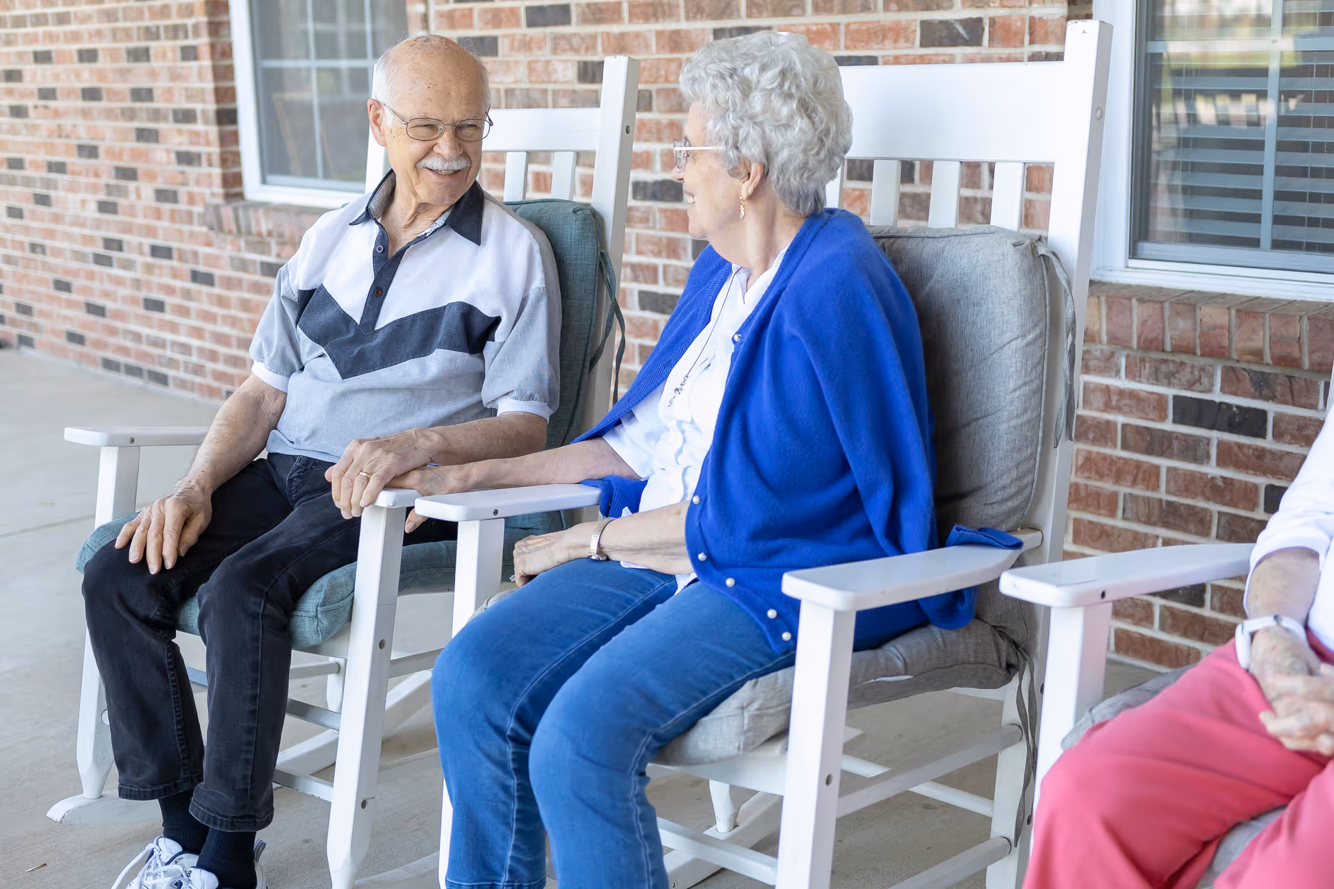 An elderly man and woman sitting on cushioned rocking chairs on a porch, holding hands and smiling at each other. The porch has a brick wall with windows in the background.