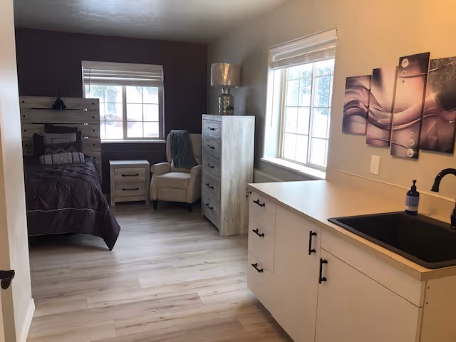 A cozy assisted living bedroom with a single bed featuring dark bedding, a wooden headboard, a nightstand, a comfortable armchair with a throw blanket, a tall dresser with a lamp on top, and a kitchenette area with white cabinets, a black sink, and a modern abstract wall art above the counter. Two large windows allow natural light to brighten the room.