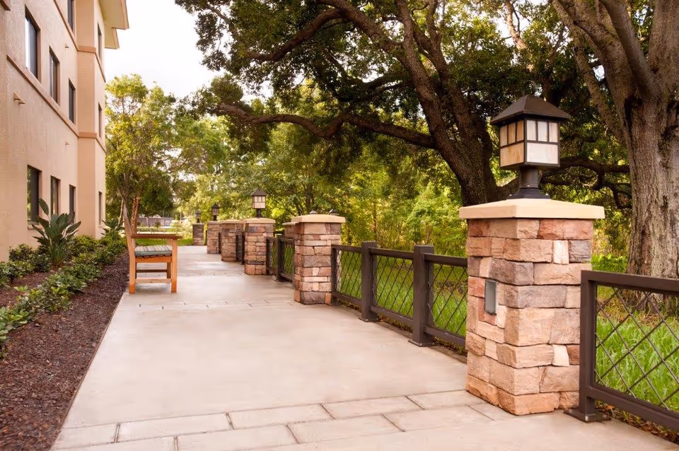 Outdoor walkway alongside a building with stone pillars topped with lantern-style lights and a wooden bench, surrounded by trees and greenery.