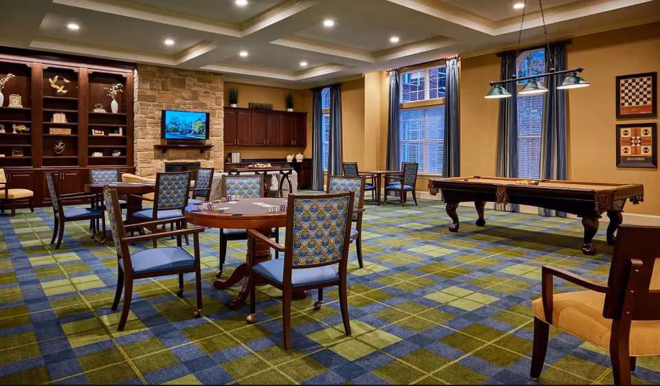 Spacious senior living common room with card tables and chairs, a pool table, fireplace with TV, built-in shelving, and large windows.