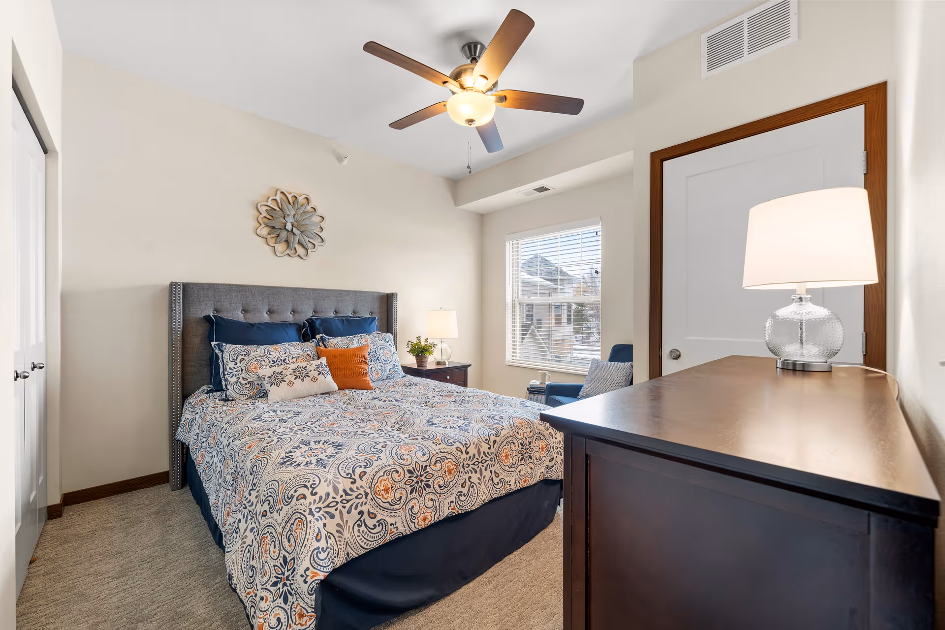 Bright furnished bedroom with a patterned bed, nightstand, dresser with lamp, ceiling fan and window.