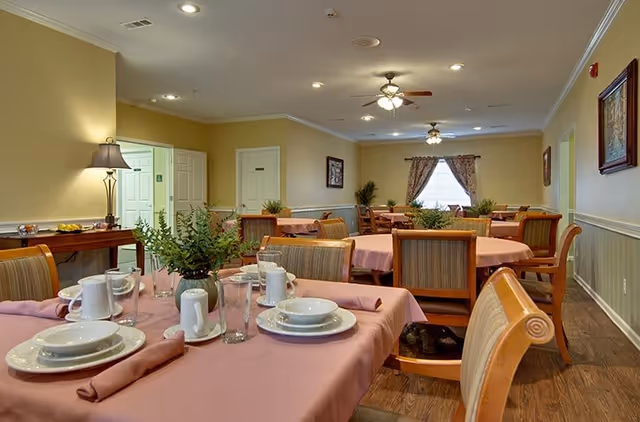 Dining room with round tables covered in pink tablecloths, place settings, chairs and potted centerpieces in a well-lit communal space.