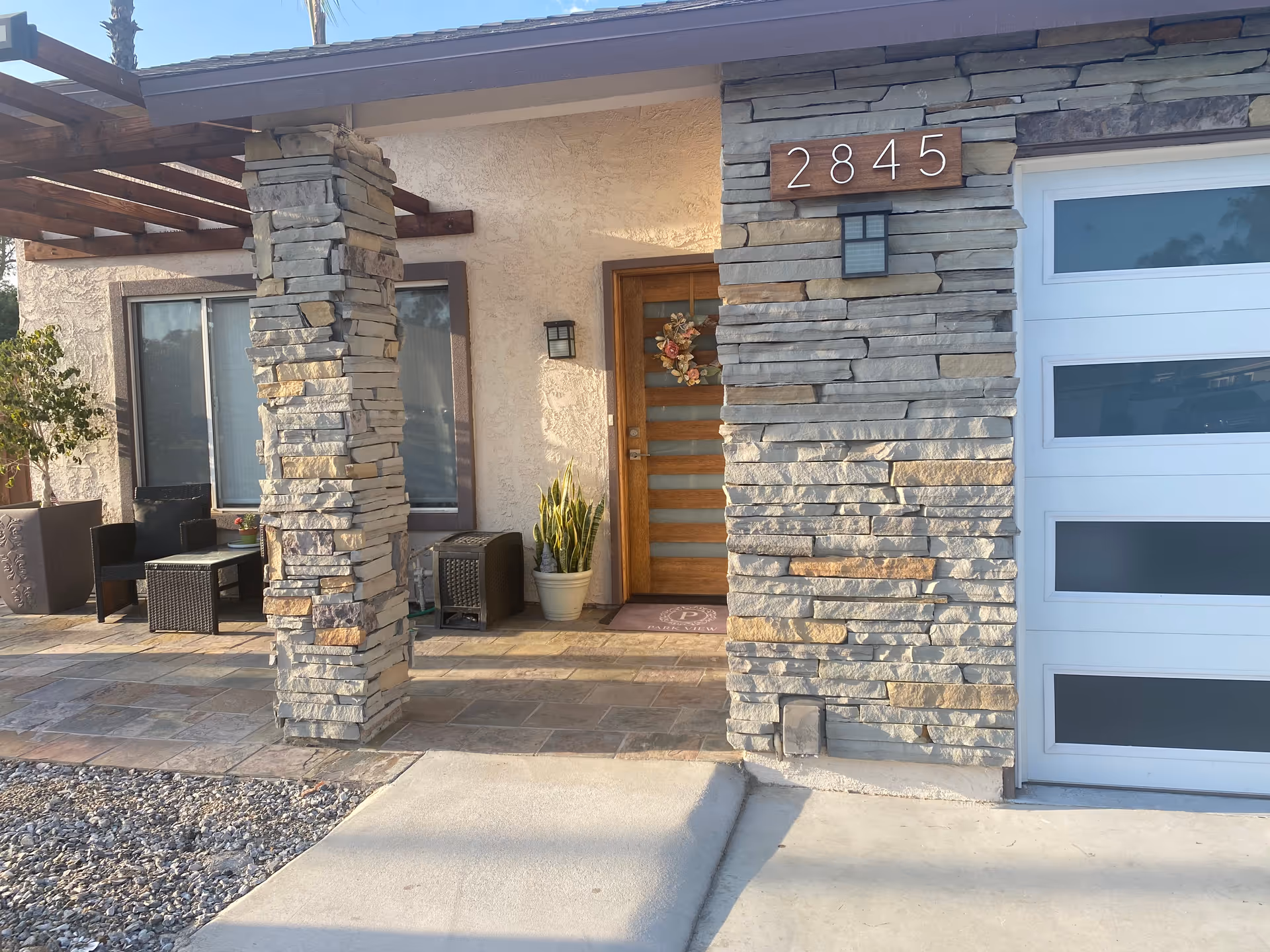 Front entrance of a residential building with stone pillars and a wooden door decorated with a wreath. The house number 2845 is displayed on a wooden plaque above a stone wall next to a modern garage door with frosted glass panels. There is outdoor seating with black wicker chairs and a table under a wooden pergola to the left of the entrance.