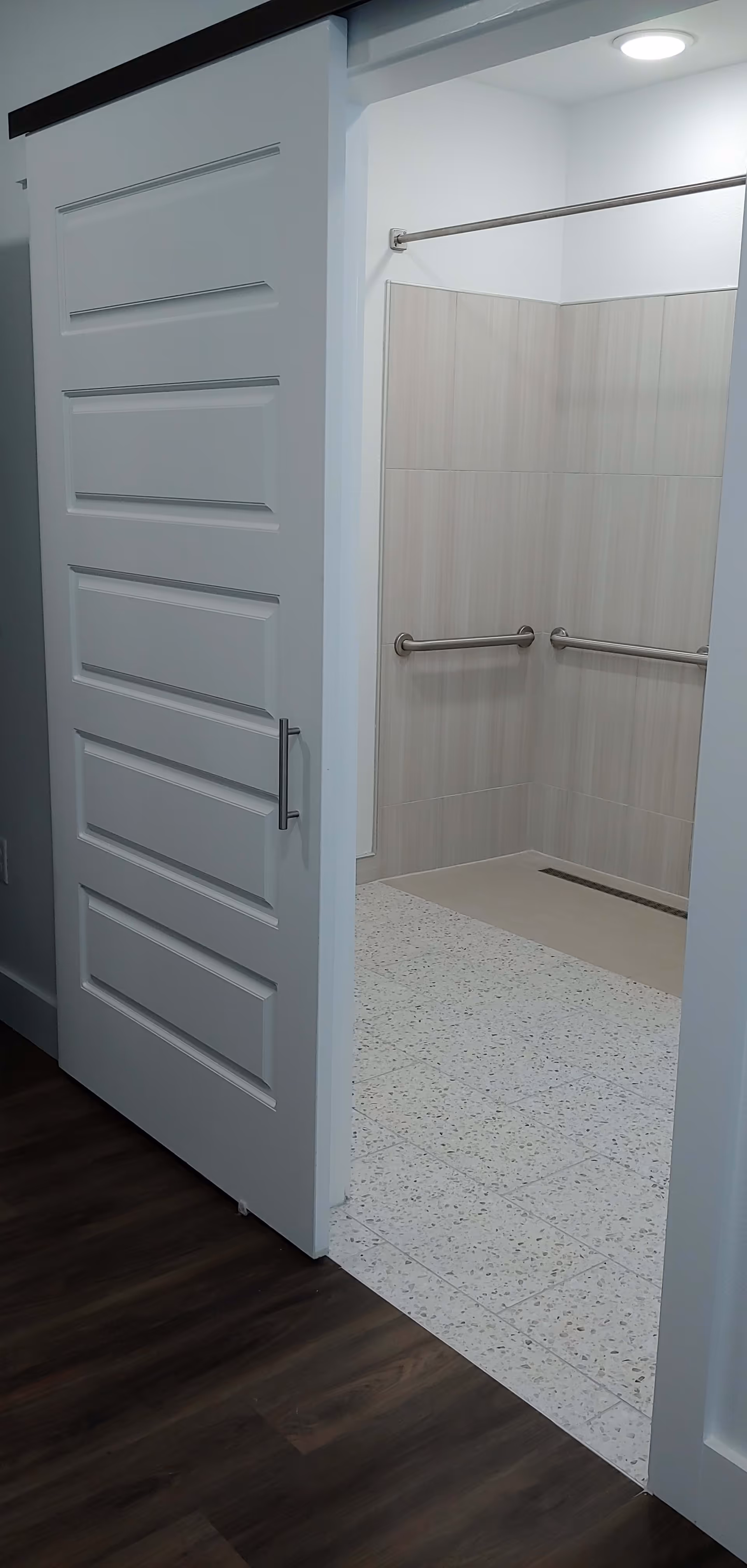 View of a bathroom entrance with a white sliding door partially open, showing a tiled shower area with two stainless steel grab bars and a shower curtain rod. The floor outside the bathroom is dark wood, and the bathroom floor is light terrazzo tile.