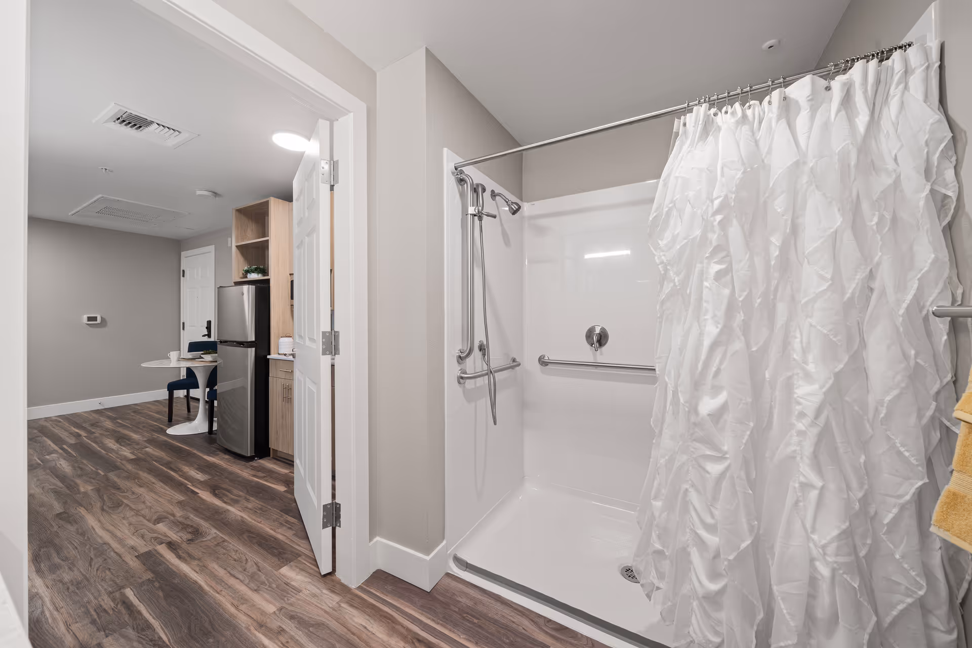 Walk-in bathroom shower with grab bars and a ruffled white curtain opening into a small kitchenette and dining area.
