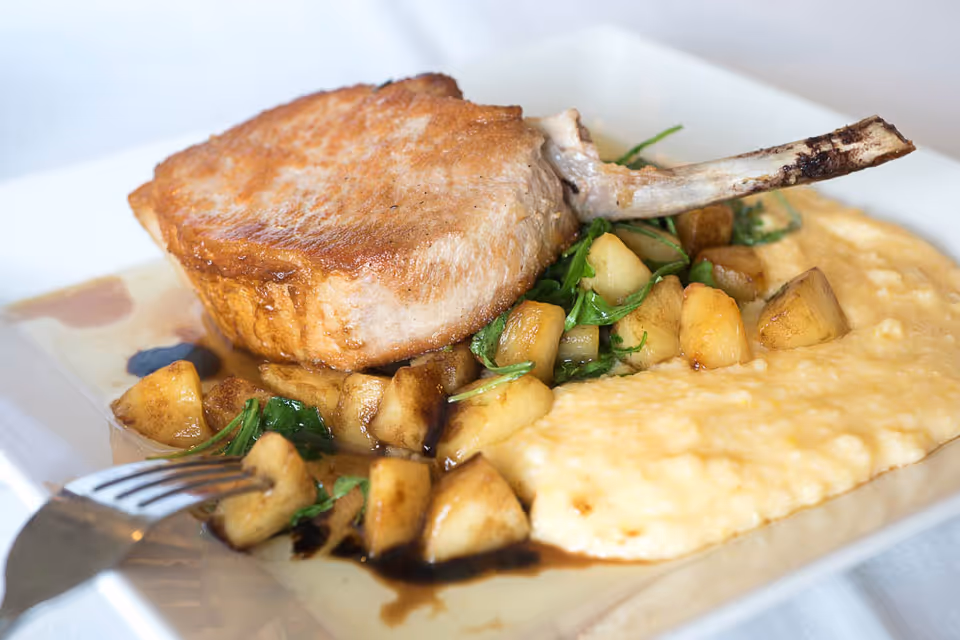 A plated pork chop served with roasted potatoes, greens and creamy mashed potatoes or polenta on a white plate.