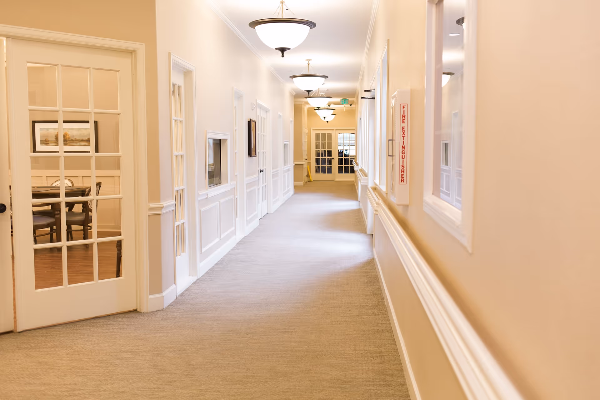 A well-lit hallway in a senior living facility with beige walls, carpeted floor, and white trim. Several closed doors line the hallway, and a glass-paneled door on the left reveals a dining area with a table and chairs. Ceiling lights are evenly spaced along the corridor, and a fire extinguisher is mounted on the right wall.