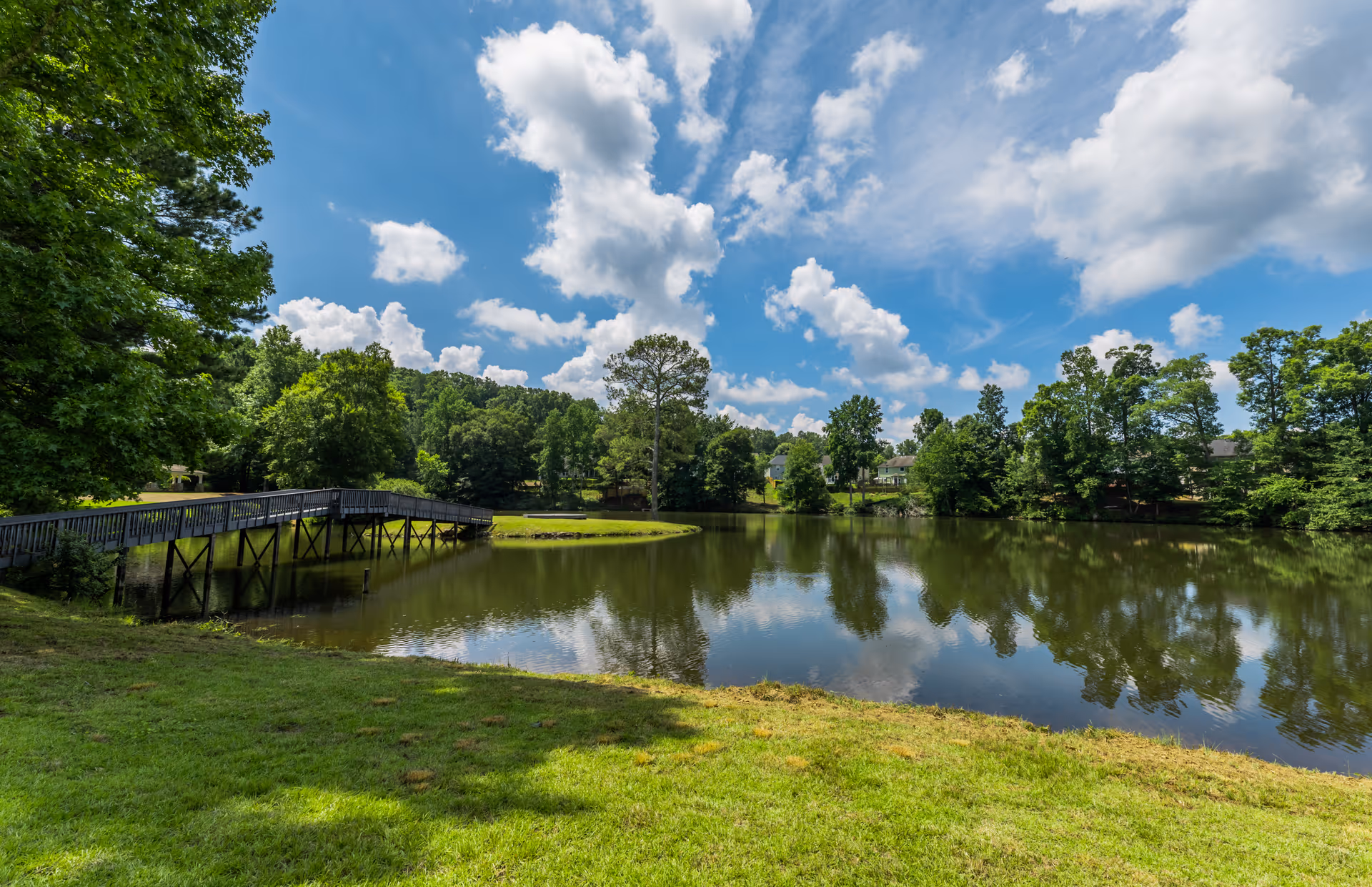 A peaceful outdoor scene featuring a small lake surrounded by green grass and trees under a partly cloudy blue sky. A wooden footbridge extends over the water to a small island with trees and shrubs.