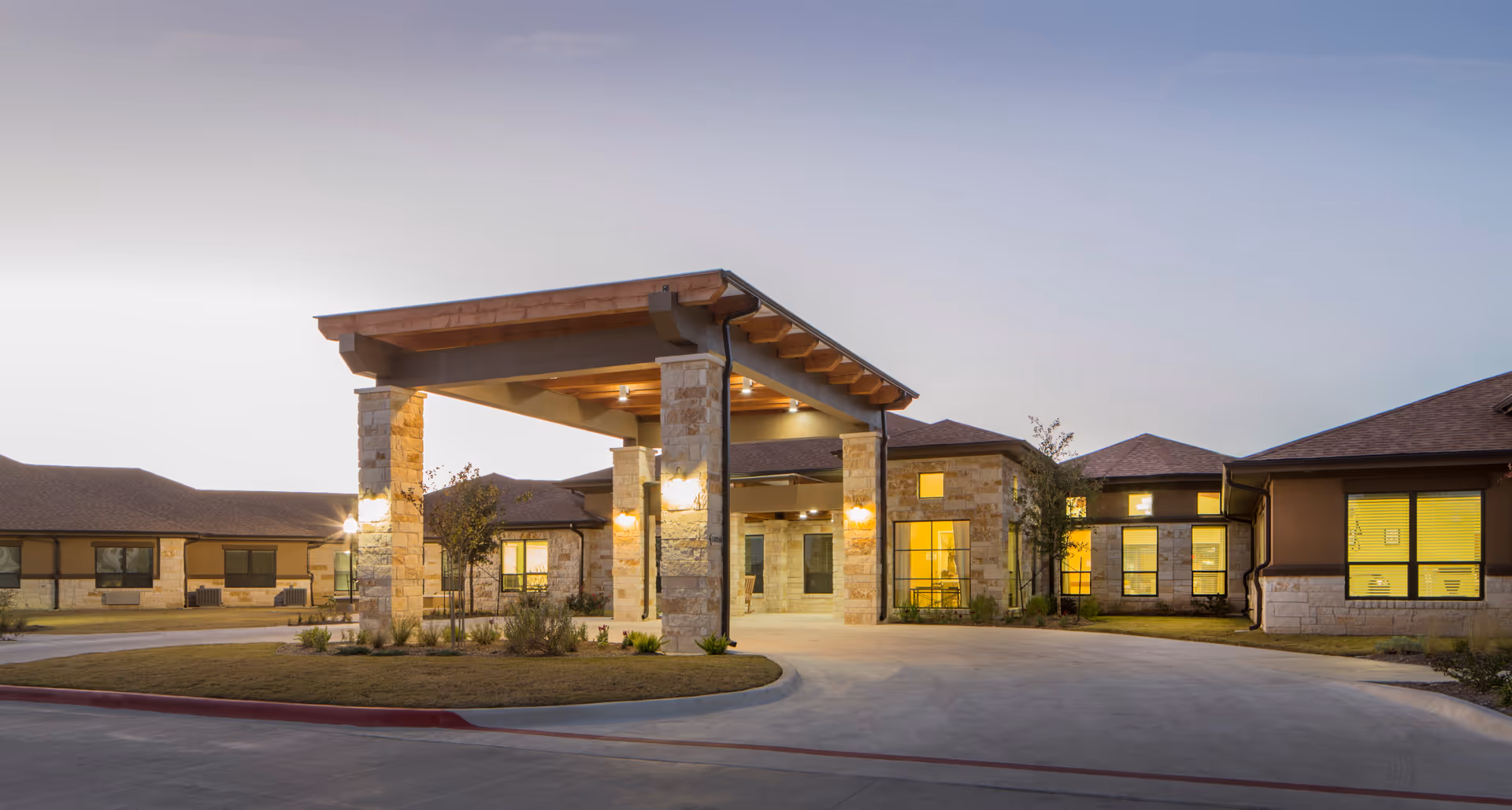 Exterior view of Park Manor Bee Cave facility at dusk, showing a single-story building with stone pillars and warm lighting under a covered entrance driveway.