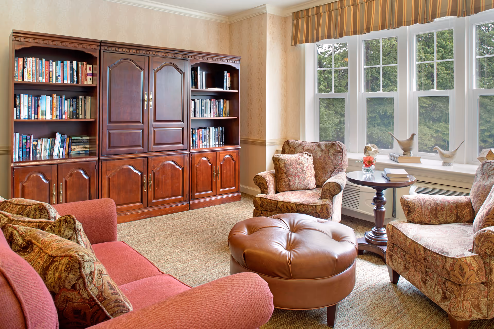 Cozy living room with upholstered chairs, a leather ottoman, a sofa, a large wooden bookcase, and a bay window overlooking trees.