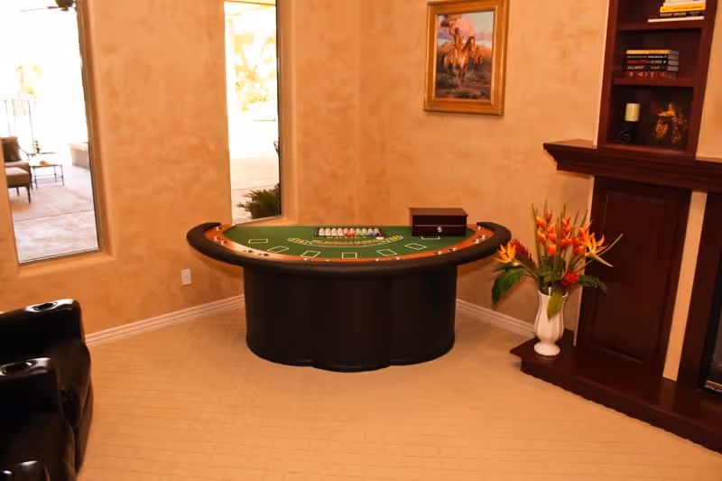A cozy interior room with beige walls and carpet, featuring a blackjack gaming table in the center, a black leather chair on the left, a wooden fireplace mantel with shelves on the right, a vase with colorful flowers on the floor, and a window showing an outdoor patio area.