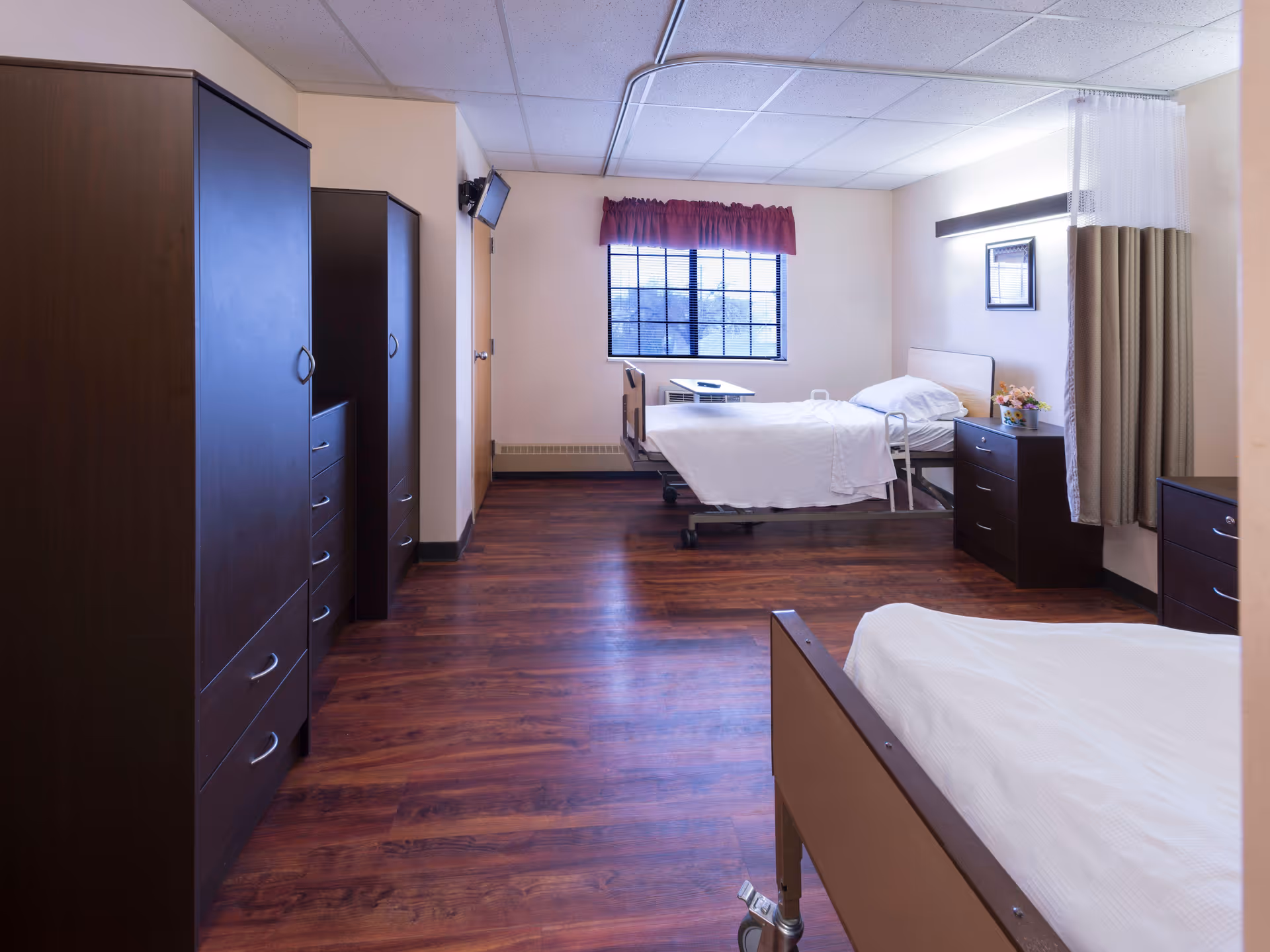 A spacious room in a healthcare facility with two hospital beds, wooden wardrobes, a window with a red valance, and a small TV mounted on the wall. The room has wooden flooring and light-colored walls, with a privacy curtain partially visible on the right side.