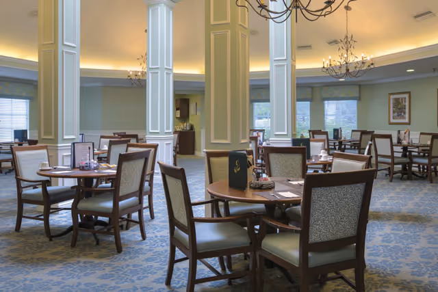 A spacious dining room with round wooden tables and cushioned chairs arranged neatly. The room features large white columns, soft lighting from chandeliers, and windows with blinds letting in natural light. The carpet has a blue floral pattern, and the walls are painted in a light green shade with framed artwork.