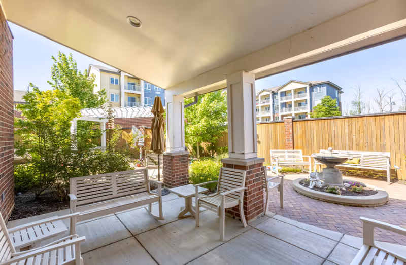 Covered outdoor patio area with white wooden benches and chairs arranged around small tables. Beyond the patio is a brick-paved courtyard featuring a circular fountain with decorative elements and additional benches. The area is surrounded by a wooden fence and greenery, with multi-story residential buildings visible in the background under a clear sky.