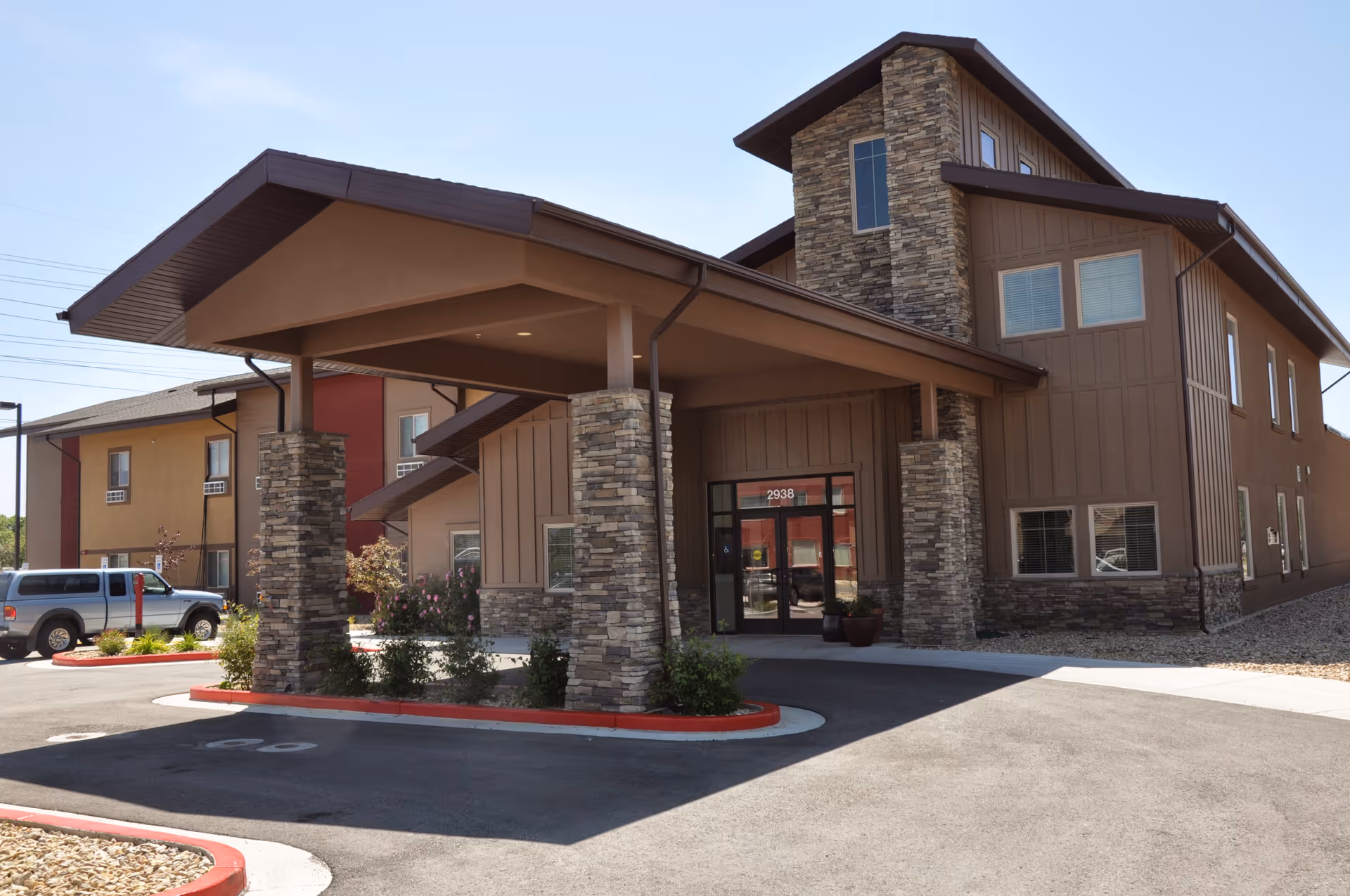 Exterior view of Tradition Assisted Living facility showing the main entrance with a covered drop-off area supported by stone pillars. The building has a brown facade with multiple windows and a clear blue sky in the background.