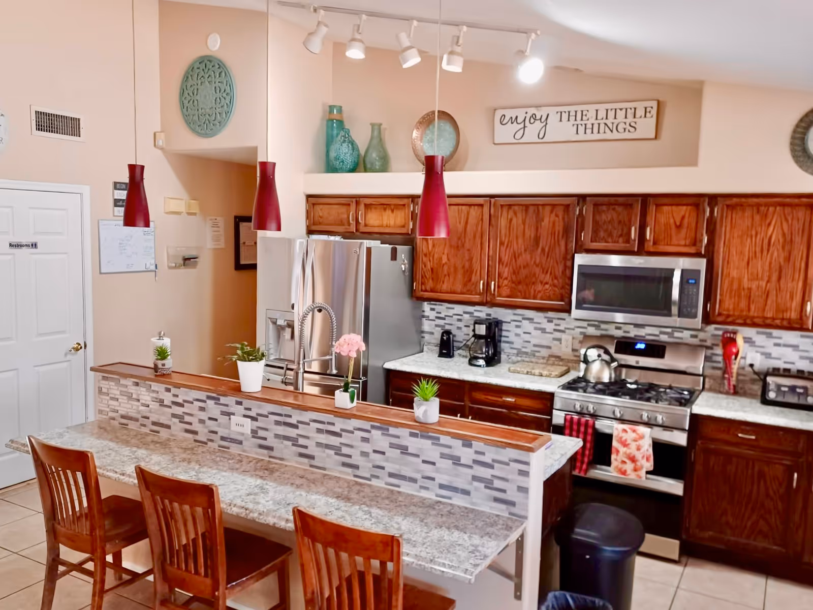 A modern kitchen with wooden cabinets, a stainless steel refrigerator, stove, and microwave. The kitchen features a granite countertop with a raised bar area and three wooden chairs. There are decorative plants and flowers on the counter, red pendant lights hanging from the ceiling, and a sign on the wall that reads 'enjoy THE LITTLE THINGS'.