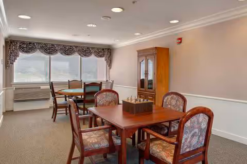 Small communal dining room with wooden tables and upholstered chairs, a china cabinet, and a large window with a decorative valance.