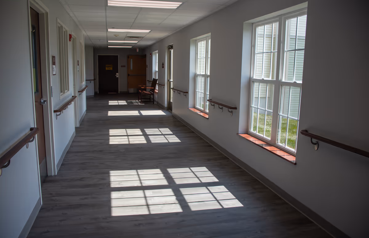 A well-lit hallway in a senior living facility with large windows on the right side casting square shadows on the wooden floor. The hallway has handrails on both sides and a wooden chair placed near the far end. Doors line the left side of the corridor.