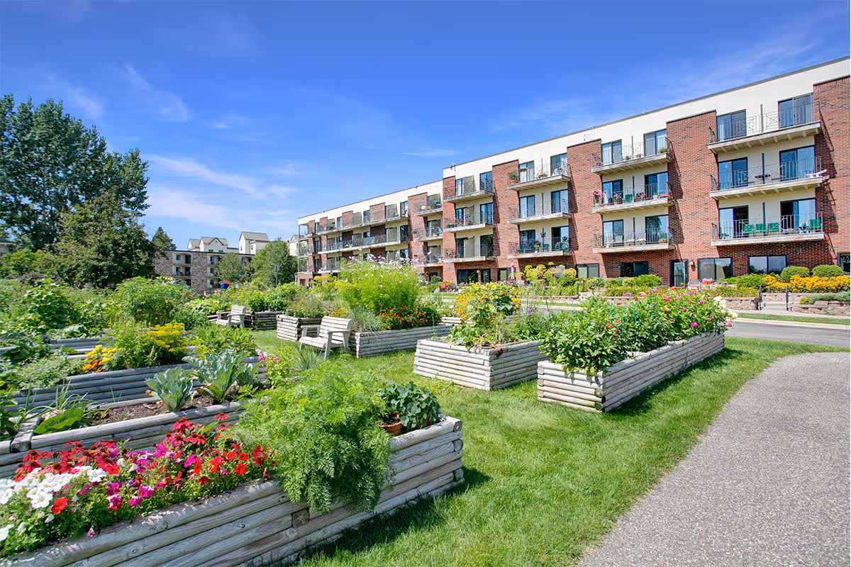 Raised wooden garden beds and benches on a lawn in front of a multi-story brick senior living building with balconies under a blue sky.