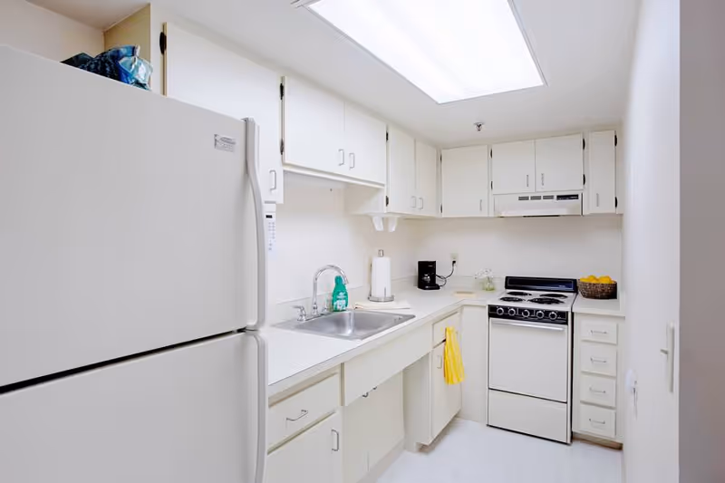 A small, clean kitchen with white cabinets, a white refrigerator, a white stove with an oven, a stainless steel sink, a coffee maker, and a basket of lemons on the counter. There is a yellow towel hanging on a cabinet handle and a bright overhead light.