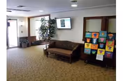 Interior view of a common area in a senior living facility with a brown couch, a potted plant, a wall-mounted television, and a display board featuring colorful artwork. There is a door with glass panels on the left side and beige carpeting on the floor.