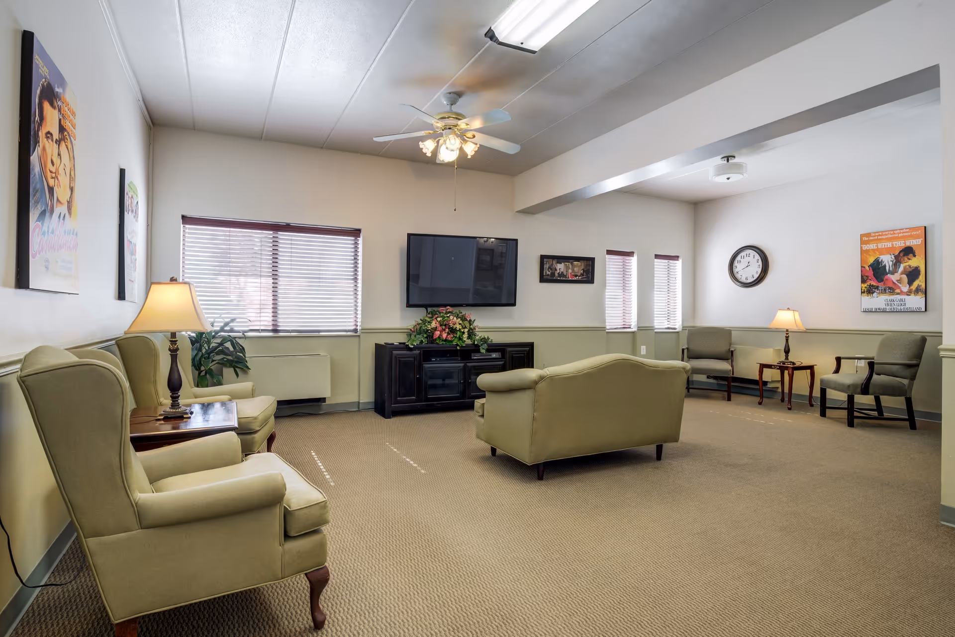 A spacious living room in an assisted living facility with beige carpet and light green walls. The room features several upholstered armchairs and a loveseat arranged around a wall-mounted flat-screen TV above a black cabinet with a floral arrangement. There are two table lamps on wooden side tables, a ceiling fan with lights, a wall clock, and framed vintage movie posters on the walls. Windows with blinds allow natural light into the room.