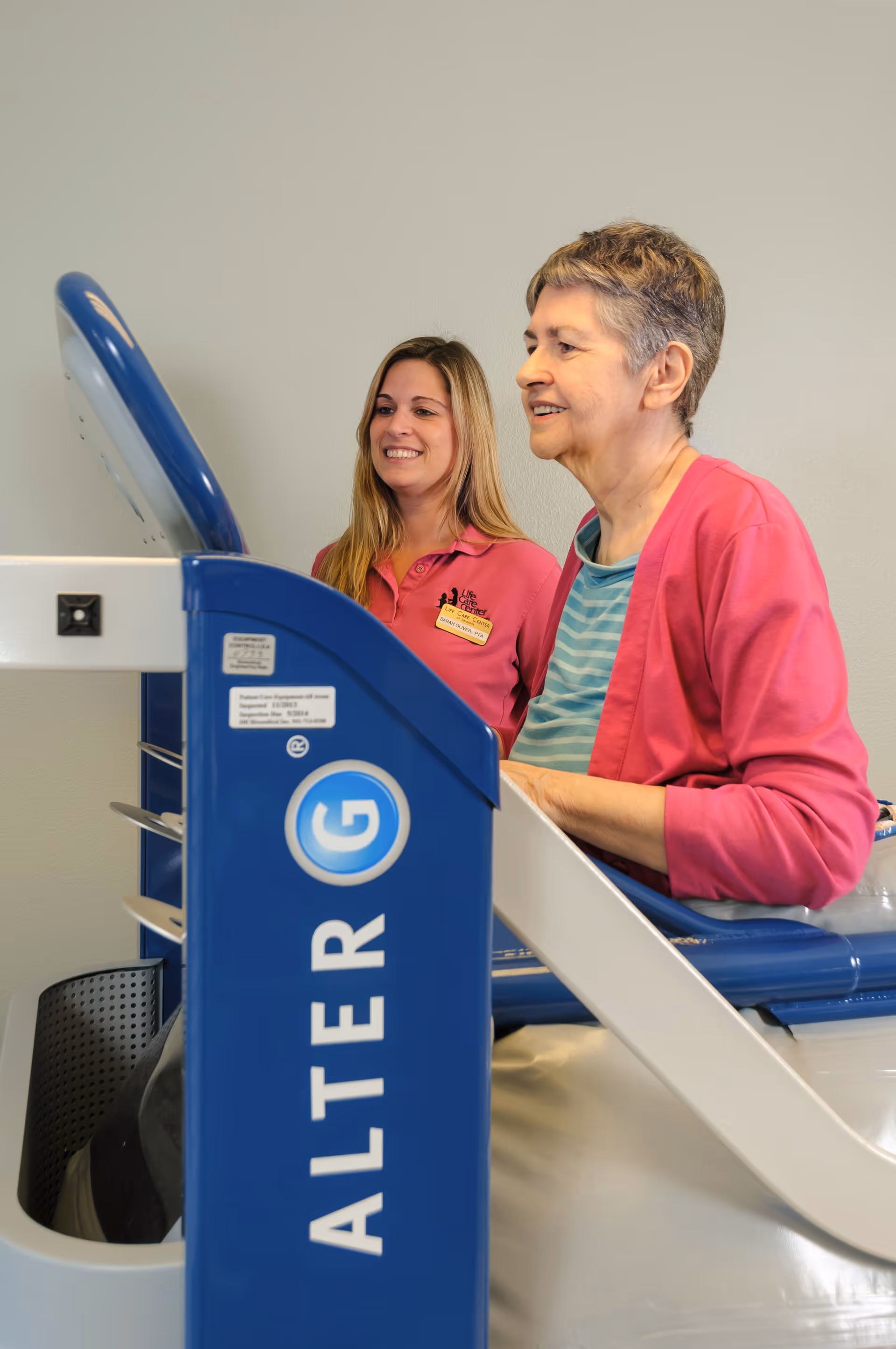 An elderly woman using an AlterG anti-gravity treadmill with the assistance of a smiling female staff member wearing a pink polo shirt in a clinical or rehabilitation setting.