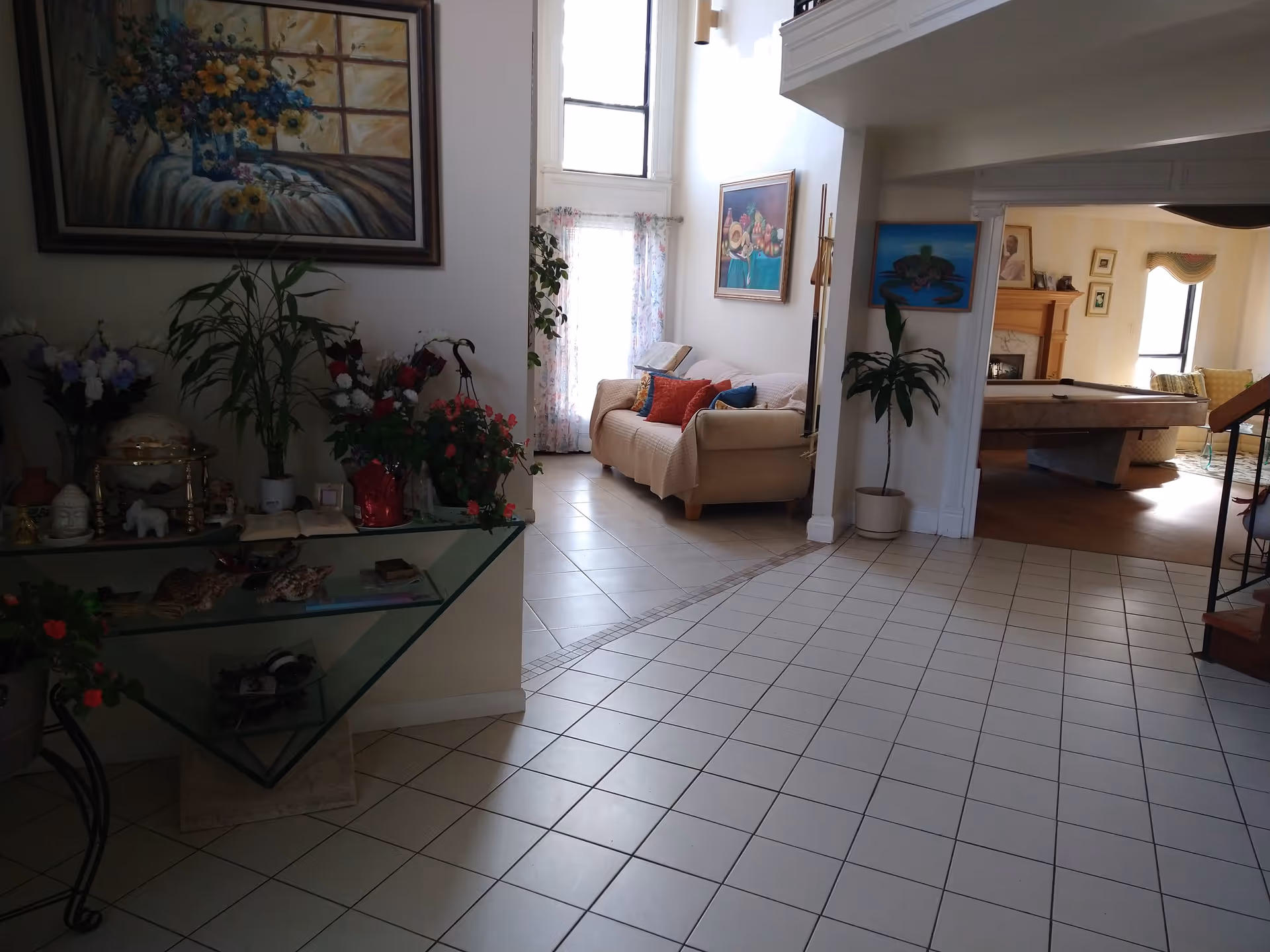 Spacious tiled interior showing an entryway leading into a living area with a sofa, plants, artwork, and a pool table in an adjacent room.