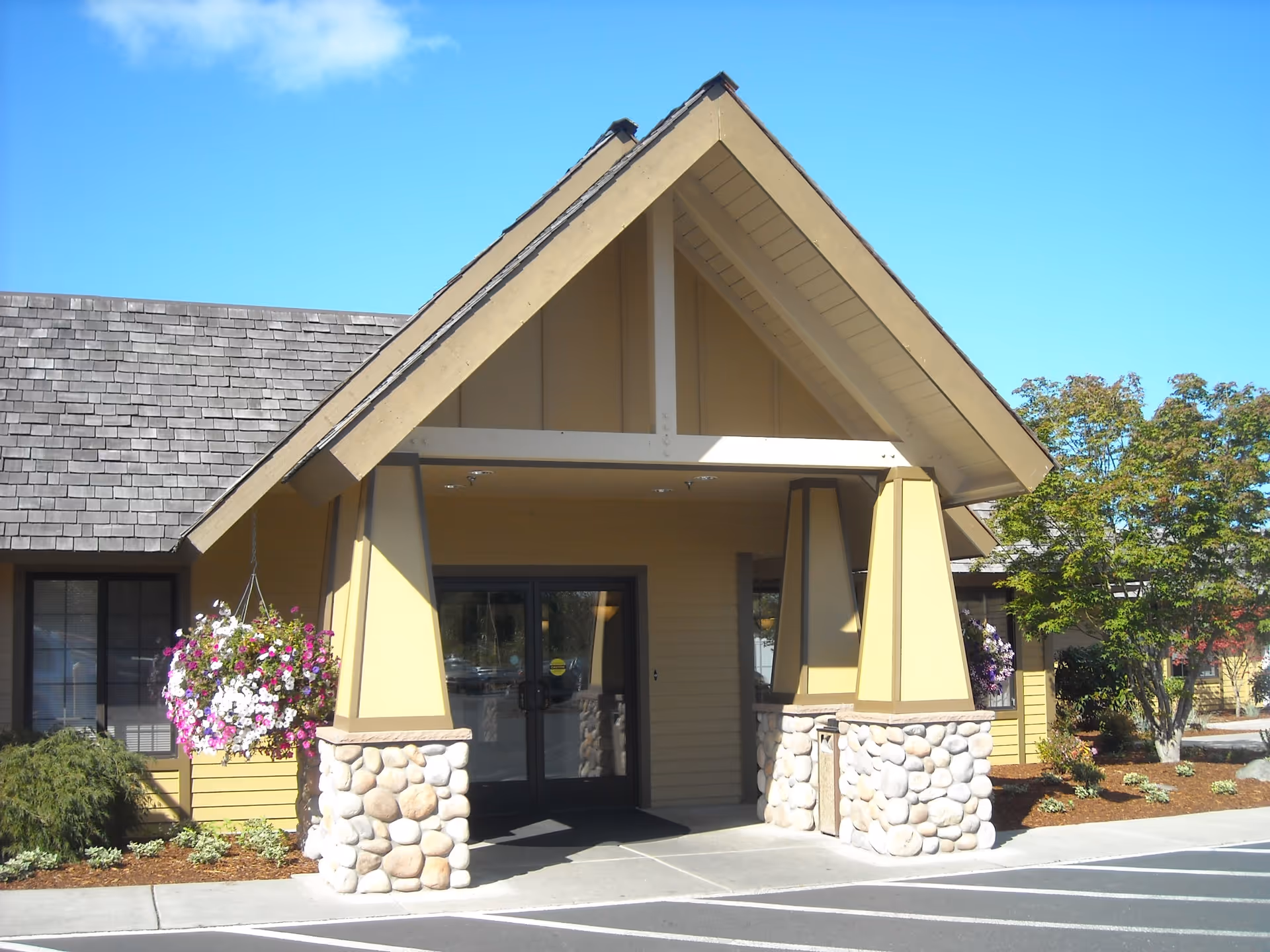 Entrance of a building with a peaked roof supported by stone and wood pillars, hanging flower baskets on either side, and a glass double door under a clear blue sky.