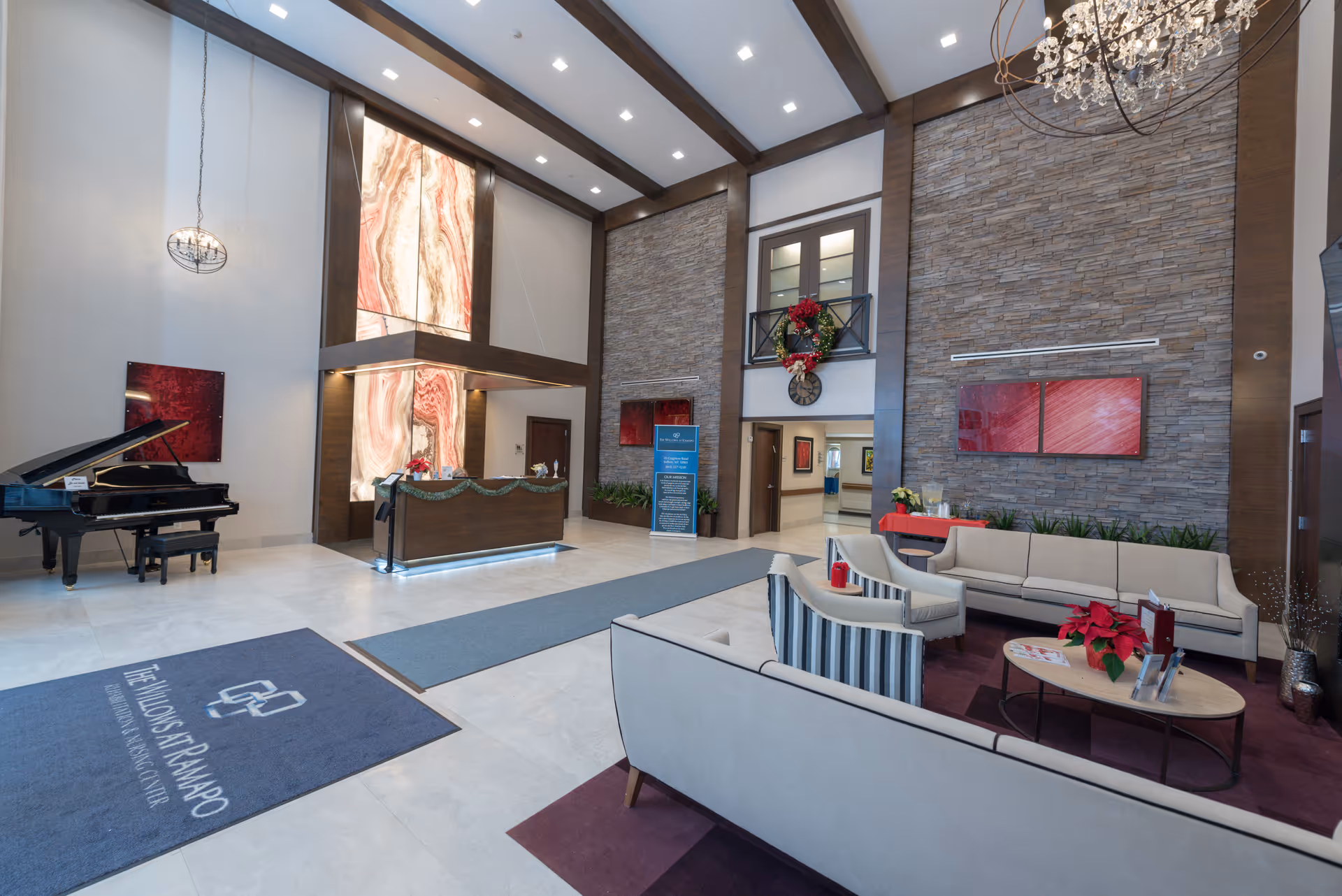 Spacious and modern lobby area of The Willows at Ramapo Rehabilitation & Nursing Center featuring a reception desk with illuminated marble panels behind it, a grand piano on the left, comfortable seating with sofas and chairs arranged around a coffee table with poinsettia plants, stone accent walls, and festive holiday decorations including a wreath above a clock.
