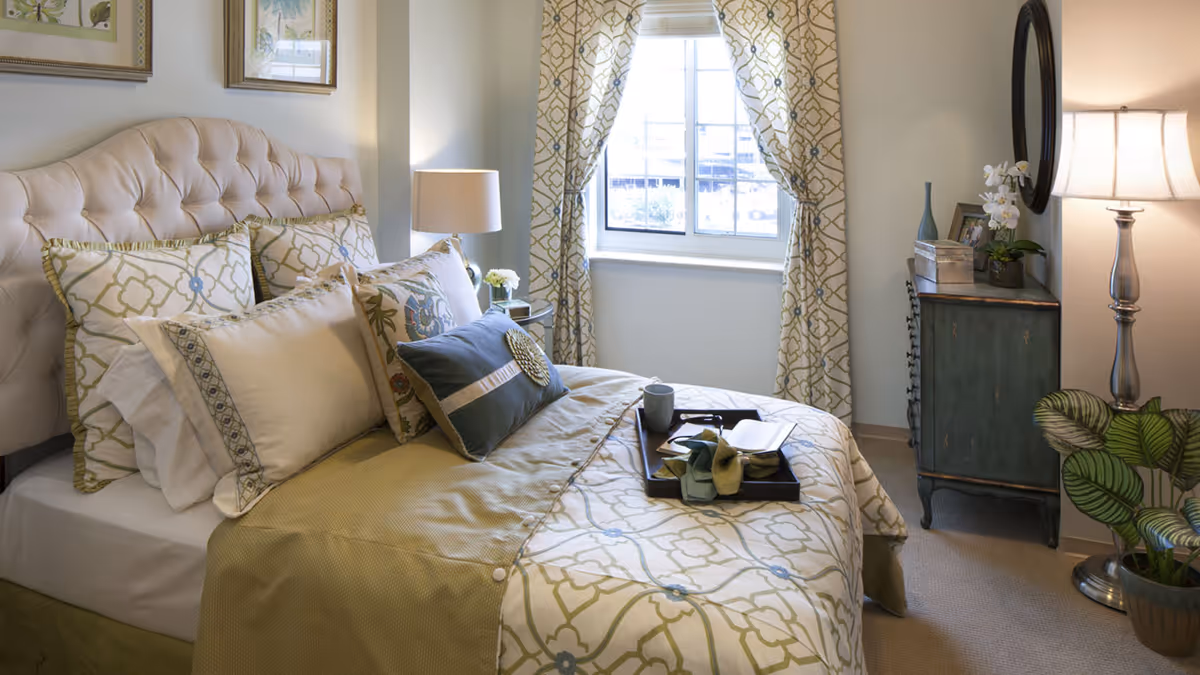 A cozy bedroom with a tufted beige headboard, a bed made with patterned pillows and a matching comforter. A tray with a cup, glasses, and a book rests on the bed. There is a window with patterned curtains letting in natural light. A small vintage-style dresser with decorative items and a round mirror is next to a tall floor lamp. A potted plant is on the floor beside the lamp.