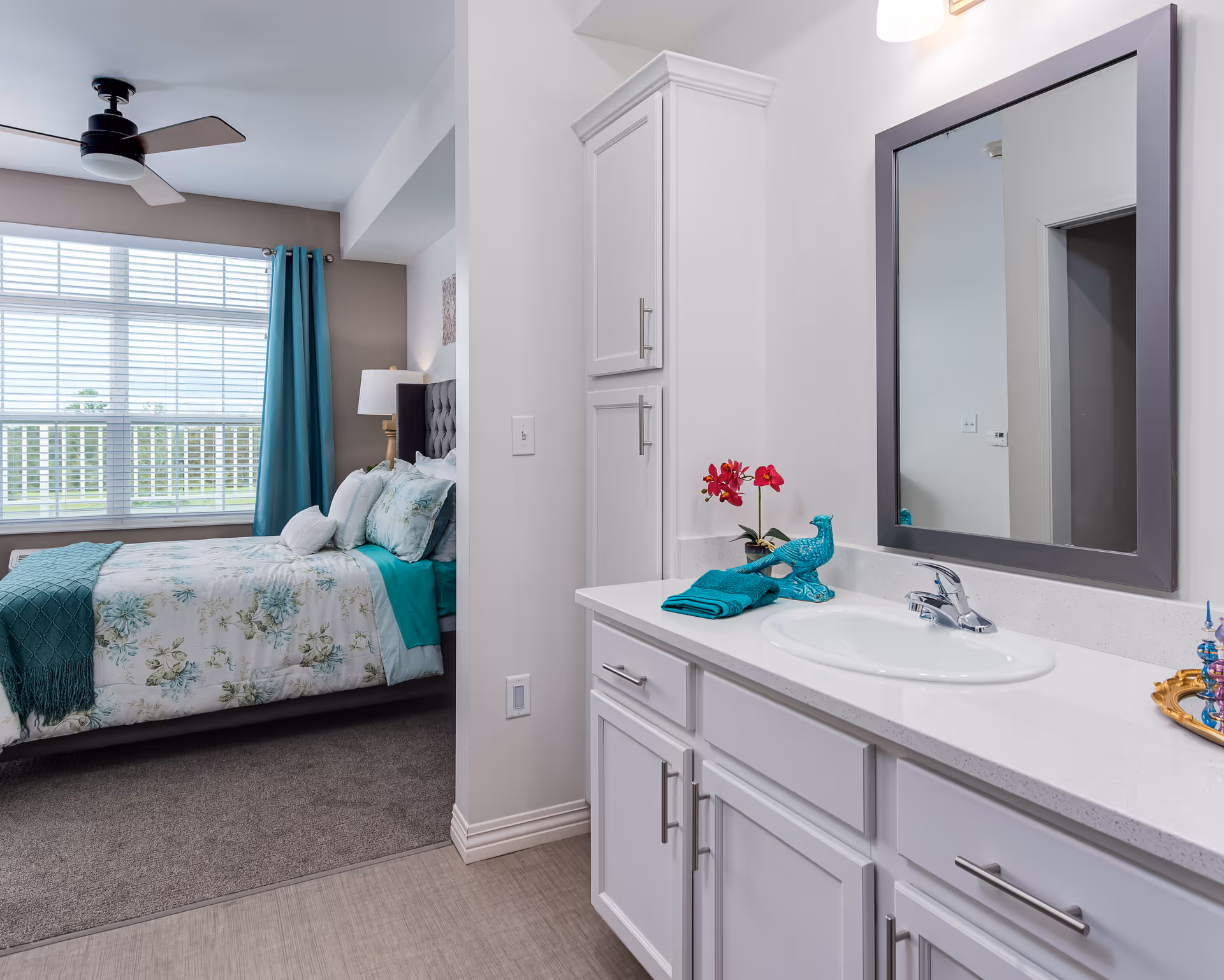 A bright bedroom with teal-accented bedding visible through an open doorway next to a white vanity sink and mirror.