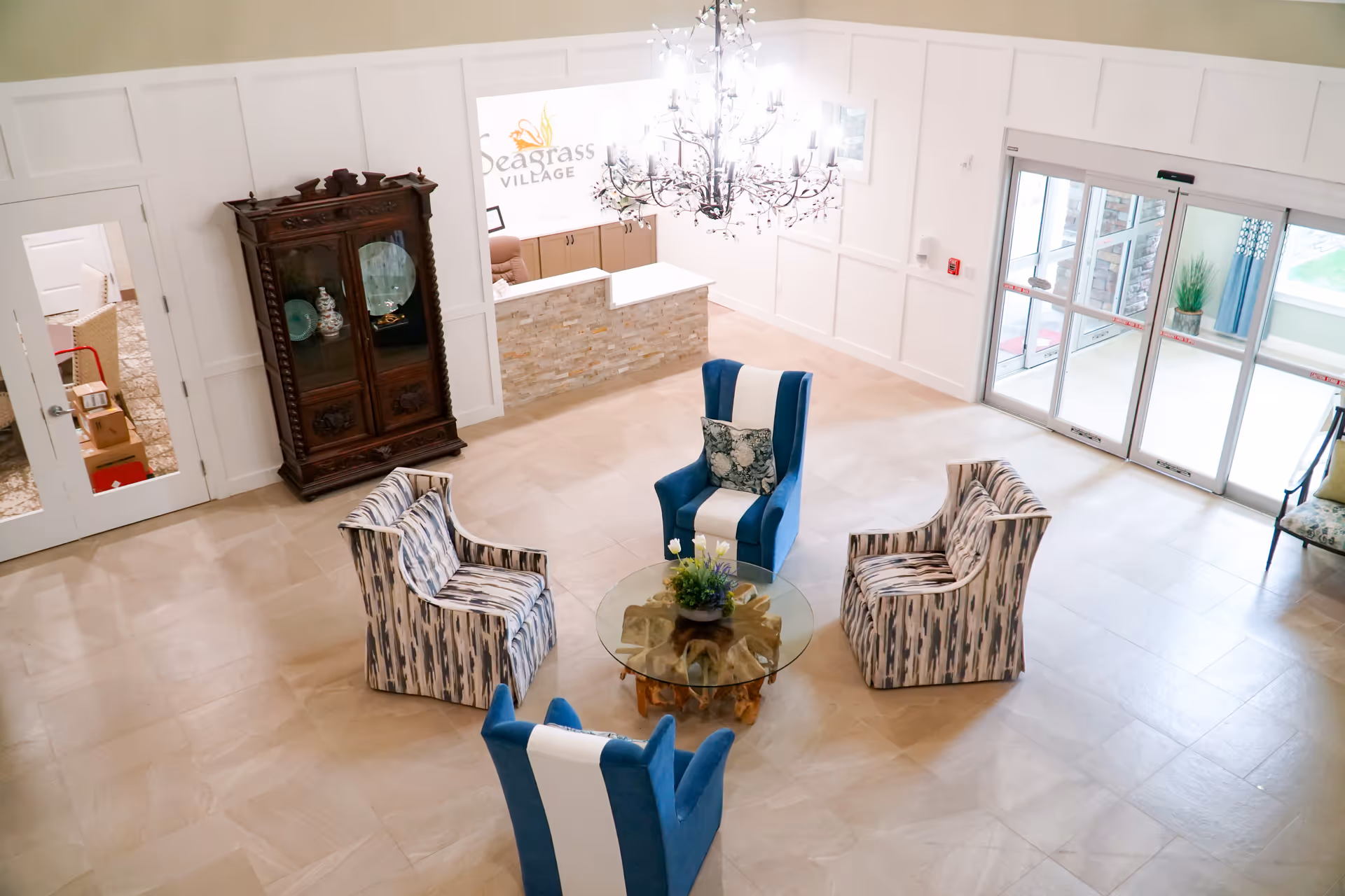 A bright and spacious lobby area with a circular seating arrangement consisting of two blue and white armchairs and two patterned armchairs around a glass coffee table with a floral centerpiece. There is a chandelier hanging from the ceiling, a wooden cabinet against the wall, a reception desk with a stone facade, and large glass sliding doors leading outside. The wall behind the reception desk displays the Seagrass Village logo.