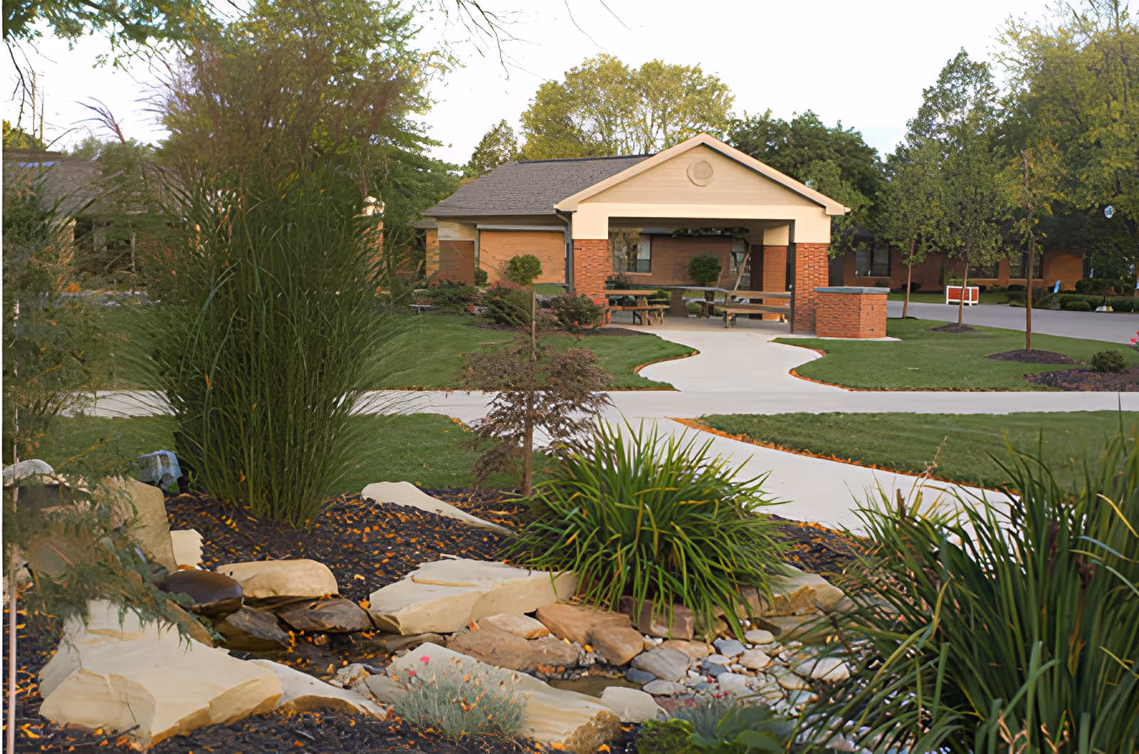 A landscaped outdoor area at Good Shepherd Home featuring a small rock garden with plants and a water feature in the foreground, a paved walkway leading to a covered seating area with benches, surrounded by green grass and trees.