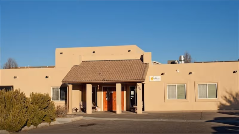 Exterior front view of a beige single-story building with a tiled roof entrance and two wooden doors, identified as BeeHive Homes of Albuquerque West, under a clear blue sky.
